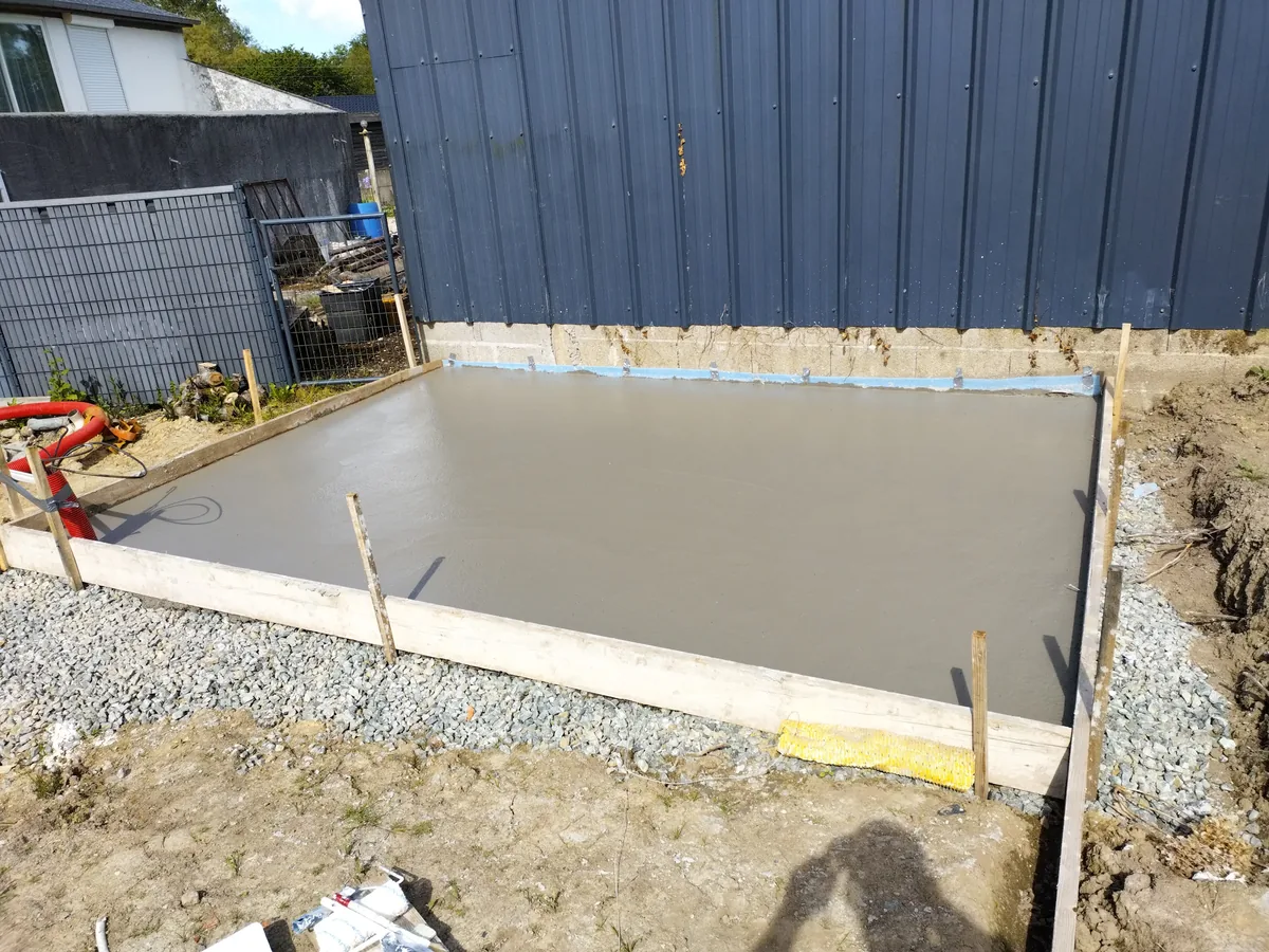 Petite dalle béton coulée dans un coffrage bois sur hérisson de gravier, le long d'un hangar bardé