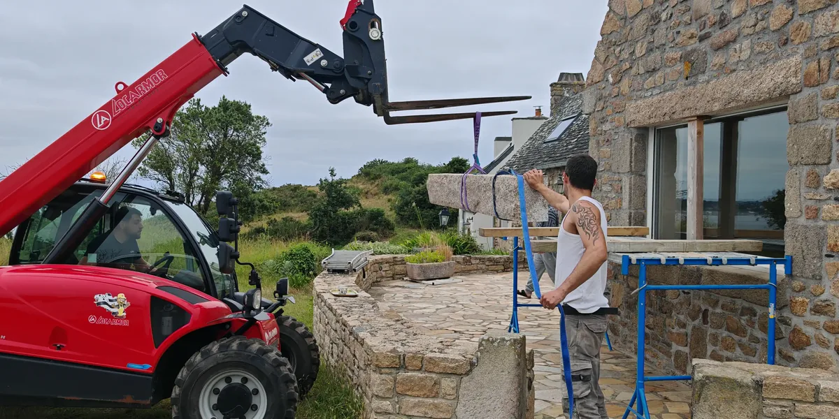 Chariot télescopique déposant un linteau en granit neuf devant une maison en pierre